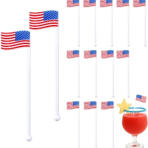 May include: A set of twelve white plastic drink stirrers, each topped with a small American flag featuring red and white stripes and a blue field with white stars. One stirrer is shown inside a glass filled with a red beverage and a star garnish.