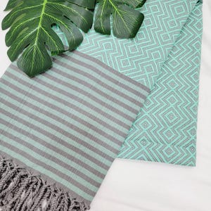 May include: A folded turquoise and gray beach towel with a geometric diamond pattern and gray stripes. The towel has a fringe on one end and is decorated with green leaves.