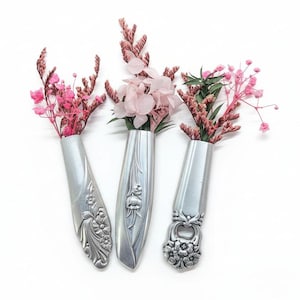 May include: Three silver spoon vases, each holding pink flowers, are arranged against a white background. The spoons have decorative floral patterns. The flowers are a mix of dried and fresh blooms, adding a touch of color and texture to the composition.