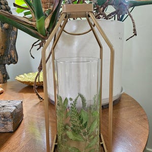 May include: A gold-colored metal lantern with a clear glass cylinder inside, decorated with green botanical designs. The lantern is on a wooden table, with a plant in the background. The lantern is a decorative home decor item.