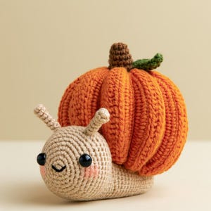 Op de afbeelding: Een gehaakt slakfiguurtje met een beige lichaam, twee voelsprieten, zwarte kraaloogjes en roze blosjes. Het slakkenhuis heeft de vorm van een ronde, geribbelde oranje pompoen met een bruin steeltje en een klein groen blaadje aan de bovenkant.