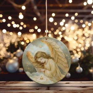 Op de afbeelding: Een rond keramisch ornament met een illustratie van een engel met grote gevederde vleugels, lang golvend haar en een aureool, met gevouwen handen in gebed boven een klein brandend kaarsje tegen een sterrenhemel, opgehangen aan een dun touwtje.