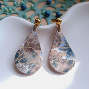 Könnte beinhalten: Ein Paar tropfenförmige Ohrringe mit floralem Muster in Blau-, Braun- und Goldtönen. Die Ohrringe haben goldfarbene Kugelstecker. Die Ohrringe werden auf einer weißen Oberfläche präsentiert. Die Ohrringe sind ca. 4 cm lang.