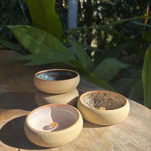 May include: Four small, handmade ceramic bowls in varying shades of tan and brown are arranged on a wooden surface. The bowls have a rounded shape with a slightly flared rim. The interior of each bowl features different colours and textures, including a speckled pattern and a teal glaze.