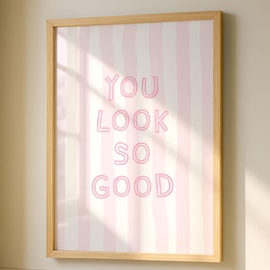 以下が含まれることがあります： ピンク色の文字で「YOU LOOK SO GOOD」と書かれた額入りのウォールアートプリント。背景にはピンクと白のストライプが垂直に並んでいます。フレームは明るい色の木材です。アートはバスルームに飾られています。