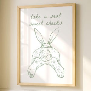 Könnte beinhalten: Ein gerahmter Druck mit hellem Holzrahmen hängt an einer Wand. Der Druck zeigt eine grüne Skizze des Hinterteils eines Kaninchens mit dem Text "take a seat sweet cheeks". Ein kleiner Holzschemel mit gefalteten weißen Handtüchern steht darunter.