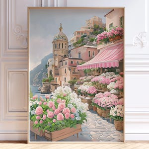 Puede incluir: Una impresión enmarcada de un pueblo costero italiano con edificios, una iglesia con cúpula y un toldo rosa. La obra de arte presenta una floristería con flores rosas y blancas en cestas y jardineras de madera. La escena se desarrolla sobre un fondo de mar y cielo azules.