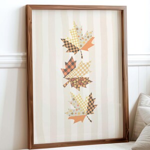 May include: A framed print with three decorative maple leaves in autumnal colours. The leaves are made from patterned fabrics, set against a striped background. The frame is made of dark wood, suitable for a living room.