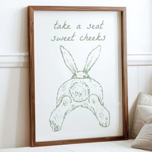 Könnte beinhalten: Ein gerahmter Druck mit einem braunen Holzrahmen. Der Druck zeigt eine grüne Illustration des Hinterteils eines Kaninchens mit dem Text "take a seat sweet cheeks" in grüner Schrift. Der Druck wird auf einem weißen Sofa angezeigt.