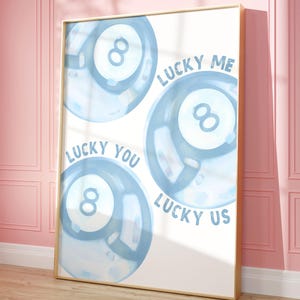 May include: A framed art print featuring three blue watercolor-style eight-ball designs. The words "LUCKY ME", "LUCKY YOU", and "LUCKY US" are written around the balls. The frame is gold, and the background is white.