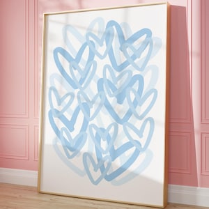 May include: A framed art print featuring a cluster of overlapping light blue hearts on a white background. The artwork is displayed in a gold frame and leans against a pink wall, creating a modern and romantic aesthetic.