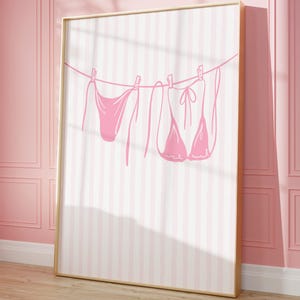 Può includere: Stampa incorniciata con lingerie rosa appesa a un filo su uno sfondo a righe rosa e bianche. L'opera d'arte si trova in una stanza con pareti rosa e pavimento in legno.