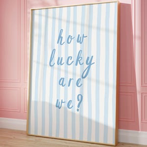 May include: A framed art print with a light blue and white striped pattern. The text "how lucky are we?" is written in a cursive font. The frame is gold, and the artwork is leaning against a pink wall.
