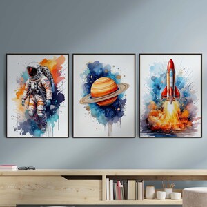 May include: Three watercolor paintings in frames, each depicting a different space-themed scene. The first shows an astronaut in a white spacesuit floating in space. The second shows a planet with rings in a blue and purple nebula. The third shows a red rocket ship launching into space.