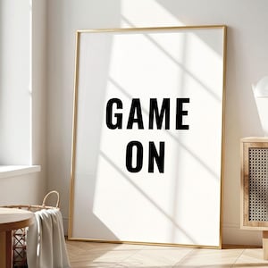 Peut inclure: Une affiche murale rectangulaire verticale dans un cadre doré fin avec le texte noir GAME ON centré sur un fond blanc. L'œuvre est présentée debout sur un sol en bois clair dans une pièce lumineuse baignée par la lumière du soleil.
