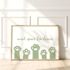 May include: A rectangular wall art print in a light wood frame featuring a white background with sage green illustrations of five cat paws at the bottom and the sage green text wash your toe beans centered above the paws.