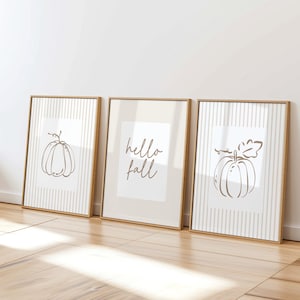Puede incluir: Tres cuadros decorativos con marcos de madera clara. Los cuadros exteriores muestran calabazas dibujadas con líneas marrones sobre un fondo blanco con rayas verticales. El cuadro central muestra el texto cursivo marrón hello fall sobre un fondo crema.