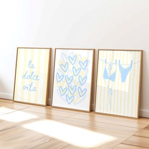 May include: Three framed wall art prints with light wood frames. The first print features yellow vertical stripes and the text la dolce vita in blue. The second shows blue heart outlines. The third displays a blue bikini on a clothesline.