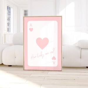May include: A framed pink and white art print featuring a playing card design. The card is the Ace of Hearts, with a large heart in the center and the text "How lucky are we?" in a script font.