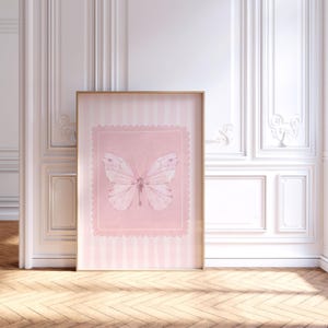 May include: A framed pink and white butterfly print. The artwork features a light pink background with a scalloped border and a watercolor-style butterfly. The frame is a light wood color, and the background has vertical stripes.