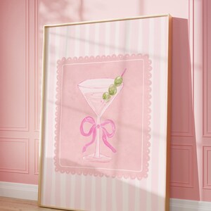 May include: A framed print featuring a pink watercolor illustration of a martini glass with olives and a bow. The artwork is set against a pink background with a scalloped border and a striped pattern. The frame is gold.