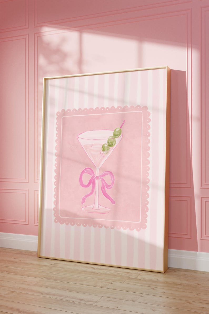 May include: A framed print featuring a pink watercolor illustration of a martini glass with olives and a bow. The artwork is set against a pink background with a scalloped border and a striped pattern. The frame is gold.