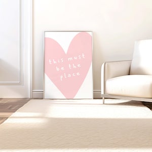 May include: A framed art print featuring a large pink heart on a white background. The words "this must be the place" are written in white script within the heart. The artwork is displayed on a white wall, leaning against a white door and a cream-colored sofa.
