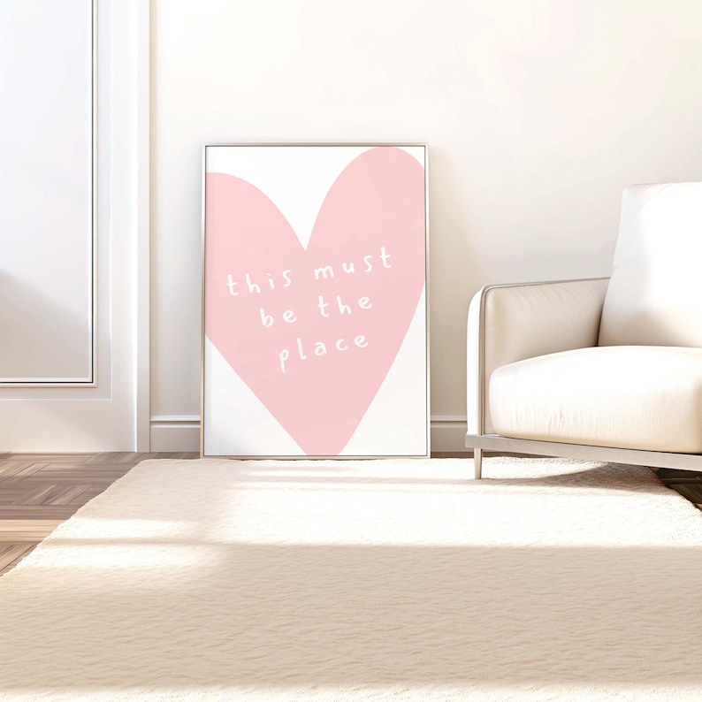 May include: A framed art print featuring a large pink heart on a white background. The words "this must be the place" are written in white script within the heart. The artwork is displayed on a white wall, leaning against a white door and a cream-colored sofa.