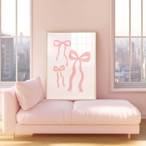 May include: A framed art print featuring three pink ribbon illustrations. The artwork is displayed above a pink chaise lounge in a room with large windows overlooking a cityscape. The room is painted in a soft pink hue.