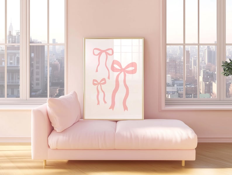 May include: A framed art print featuring three pink ribbon illustrations. The artwork is displayed above a pink chaise lounge in a room with large windows overlooking a cityscape. The room is painted in a soft pink hue.