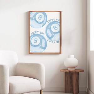 May include: A framed print featuring three blue watercolor-style eight-ball designs. Text on the print reads "Lucky Me," "Lucky You," and "Lucky Us." The artwork is displayed in a room with a white chair and a wooden side table.