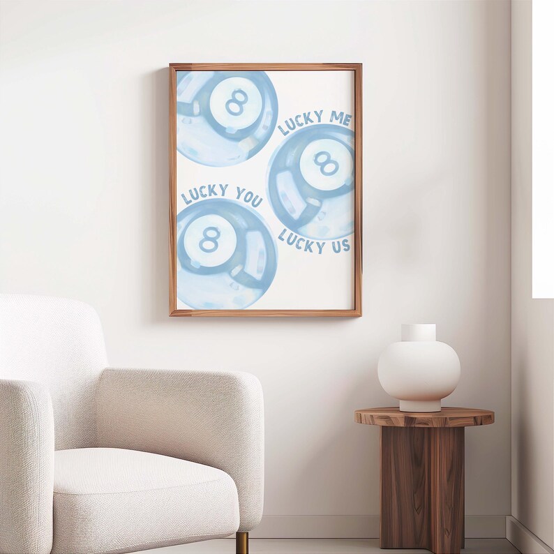 May include: A framed print featuring three blue watercolor-style eight-ball designs. Text on the print reads "Lucky Me," "Lucky You," and "Lucky Us." The artwork is displayed in a room with a white chair and a wooden side table.