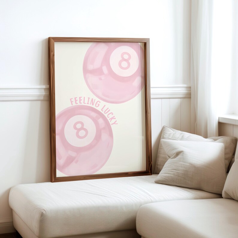 May include: A framed art print featuring two pink watercolor-style eight balls with the text "Feeling Lucky." The artwork has a cream-colored background and is set in a dark brown wooden frame. The print is displayed on a white couch.