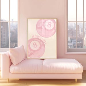 May include: A framed print featuring two pink eight balls with the text "Feeling Lucky" in a light pink room. A pink chaise lounge with a pillow is in front of the windows.
