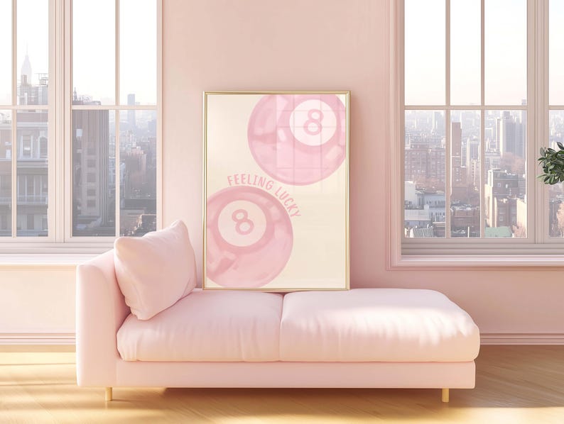 May include: A framed print featuring two pink eight balls with the text "Feeling Lucky" in a light pink room. A pink chaise lounge with a pillow is in front of the windows.
