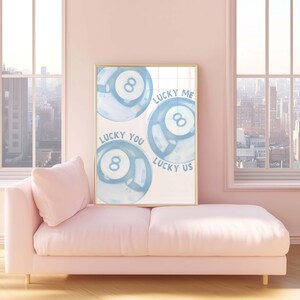 May include: A framed art print featuring three blue watercolor-style eight balls. The text on the print reads "Lucky Me," "Lucky You," and "Lucky Us." The print is displayed in a room with a pink sofa and a city view.