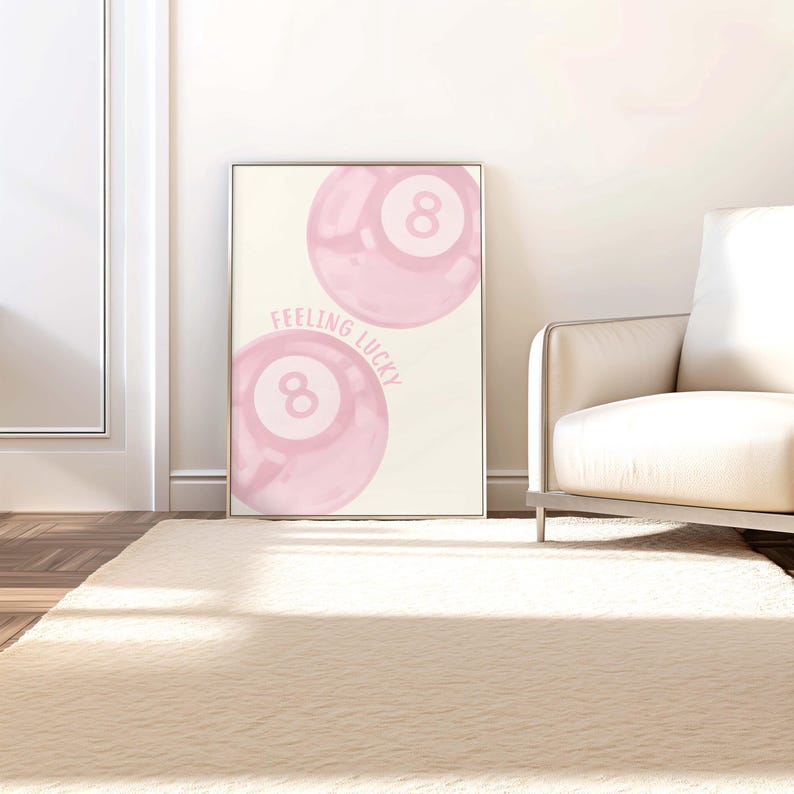 May include: A framed print featuring two pink eight balls with the text "Feeling Lucky." The artwork has a soft, watercolor style. The print is displayed in a silver frame, leaning against a white wall.