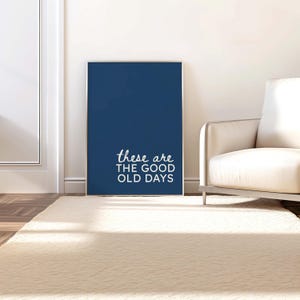 May include: A framed navy blue art print with the text "these are the good old days" in white script. The artwork is displayed on a white wall, leaning against it, with a cream-colored rug in front.