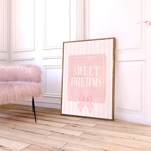 May include: A framed pink and white striped print with the words "SWEET DREAMS" and a bow illustration. A fluffy pink chair with black legs sits to the left. The room has white paneling and light wood floors.