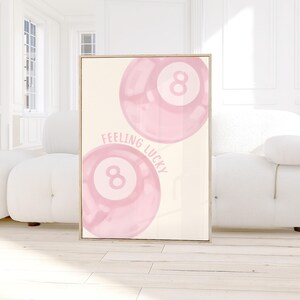 May include: A framed print featuring two pink eight balls with the text "FEELING LUCKY" in a light pink font. The artwork has a cream-colored background and a wooden frame. The image is displayed in a bright, modern room.