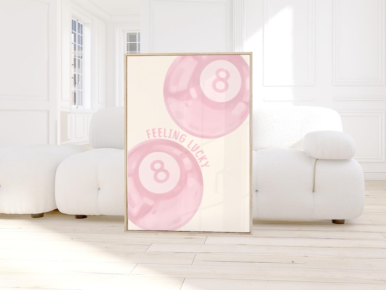 May include: A framed print featuring two pink eight balls with the text "FEELING LUCKY" in a light pink font. The artwork has a cream-colored background and a wooden frame. The image is displayed in a bright, modern room.