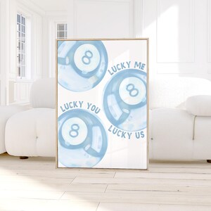 May include: A framed print featuring three light blue watercolor-style 8-ball illustrations. The words "LUCKY ME", "LUCKY YOU", and "LUCKY US" are written around the balls. The artwork has a light wooden frame.