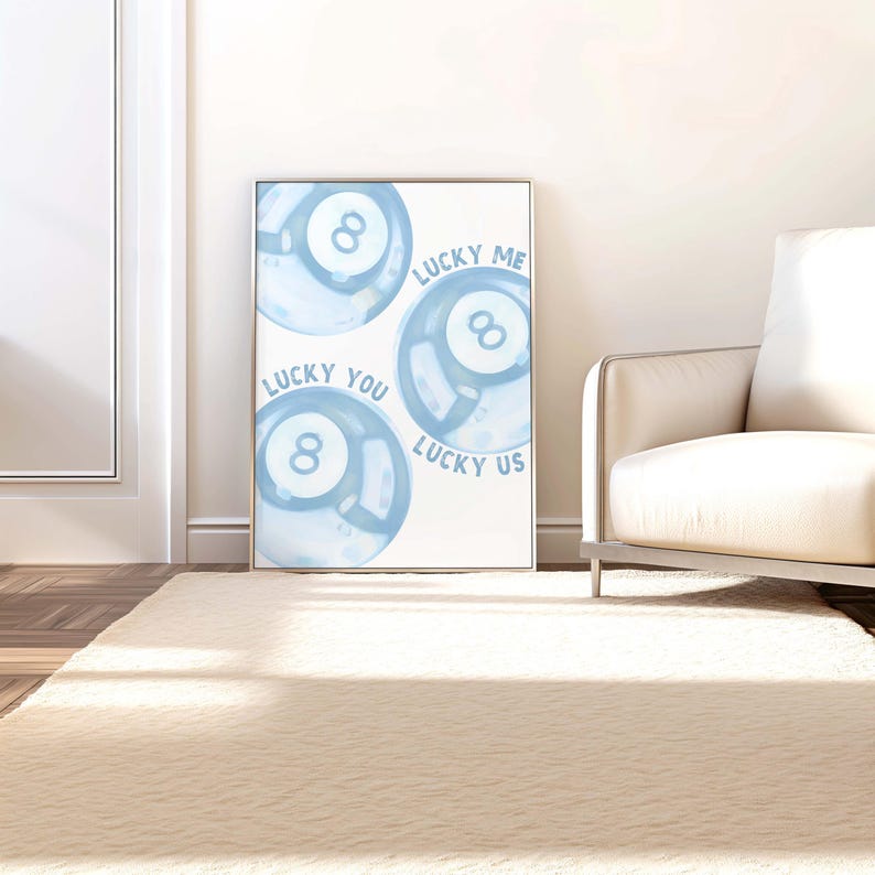 May include: A framed print featuring three light blue watercolor-style 8-ball designs. The text "LUCKY ME", "LUCKY YOU", and "LUCKY US" is written around the balls. The artwork is displayed in a bright room with a white wall and a beige rug.