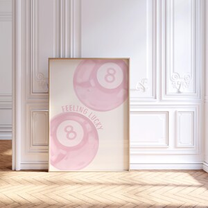 May include: A framed art print featuring two pink watercolor-style eight balls with the text "Feeling Lucky". The artwork has a light beige background and is displayed in a room with white walls and a wooden floor.