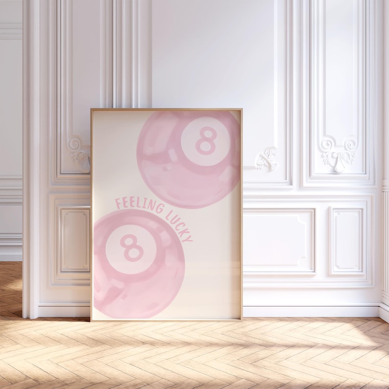 May include: A framed art print featuring two pink watercolor-style eight balls with the text "Feeling Lucky". The artwork has a light beige background and is displayed in a room with white walls and a wooden floor.
