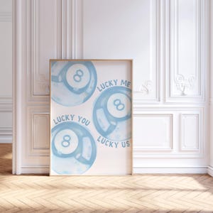 May include: A framed art print featuring three light blue 8-ball designs with the phrases "Lucky You", "Lucky Me", and "Lucky Us" in a handwritten font. The artwork is displayed in a room with white walls and a wooden floor.