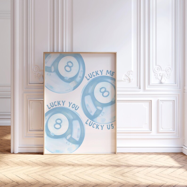 May include: A framed art print featuring three light blue 8-ball designs with the phrases "Lucky You", "Lucky Me", and "Lucky Us" in a handwritten font. The artwork is displayed in a room with white walls and a wooden floor.