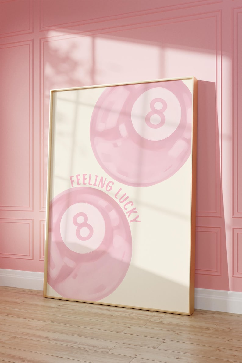 May include: A framed print featuring two pink eight-ball illustrations with the text "Feeling Lucky." The artwork has a light beige background and a gold frame. The print is leaning against a pink wall and wooden floor.