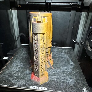 May include: A 3D printed tower-shaped mug with a textured surface and intricate details. The mug is printed in a light brown colour and has a red base. The mug is sitting on a black surface with the word "CREALITY" printed on it.