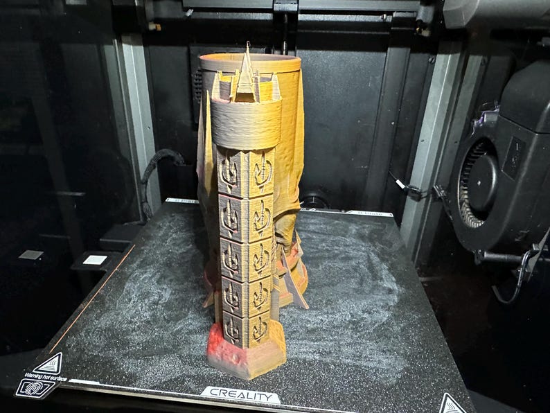 May include: A 3D printed tower-shaped mug with a textured surface and intricate details. The mug is printed in a light brown colour and has a red base. The mug is sitting on a black surface with the word "CREALITY" printed on it.